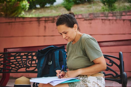Woman sitting on a bench with a backpack, attentively writing on papers. An outdoor environment, illustrating studying, focus, and diligent work in a serene setting.
