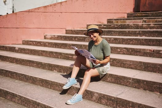 A cheerful woman sitting on outdoor steps, relaxing while reading a magazine and enjoying the sunny weather. Dressed casually, wearing a hat, this represents leisure, joy, and relaxation.