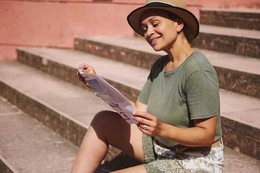 A cheerful woman enjoys a sunny day seated on stairs, wearing light summer attire, and reading a pamphlet. Bright environment emphasizing leisure and happiness.