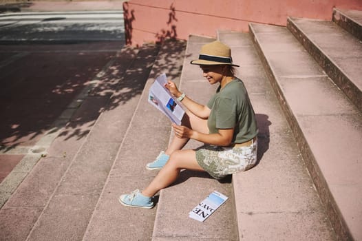 A woman reads a booklet while sitting on concrete steps in sunshine. She wears a summer outfit including a hat and sneakers. A travel brochure is on a step nearby.