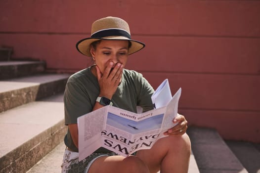 A woman in casual attire, sitting on stone steps outdoors, reads a newspaper with a surprised and emotional expression, indicating unexpected or impactful news.