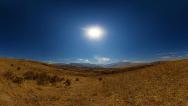 A wide panoramic view of arid, yellow-brown rolling hills under a clear blue sky with the sun shining.
