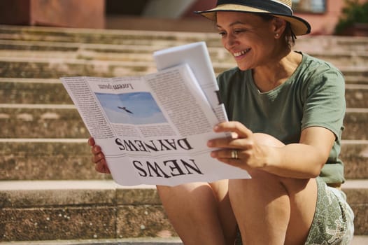 A joyful middle-aged woman enjoys reading a newspaper while sitting on outdoor steps under the sun, showcasing leisure and happiness.