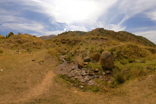 A small stream emerges from the ground in a hilly, semi-arid landscape. Green vegetation contrasts with dry slopes. Large boulder near water.