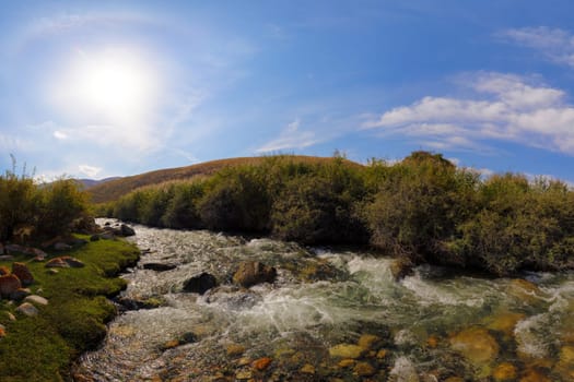 Fast-flowing mountain river cascades over rocks, creating white water rapids. Lush green banks under a bright sun and blue sky with clouds.