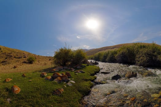 Fast-flowing mountain river cascades over rocks, creating white water rapids. Lush green banks under a bright sun and blue sky with clouds.