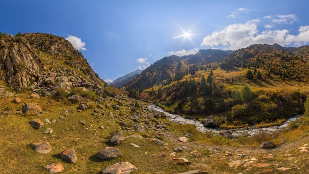 View across Zhylamysh Gorge showing a river, steep rocky cliffs, and slopes covered in green conifers and vibrant autumn foliage under a sunny sky.