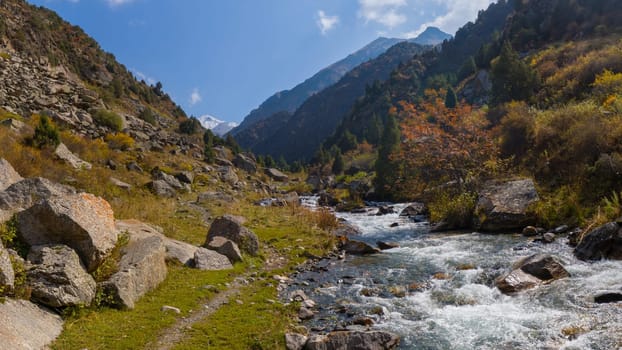 A wide shot captures a vibrant mountain river cascading over rocks, flanked by steep, rocky banks covered with patches of green and yellow vegetation.