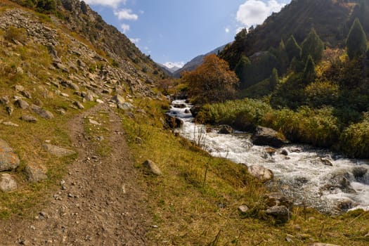 A fast-flowing mountain river with white rapids carves through a valley, bordered by a dirt hiking path.