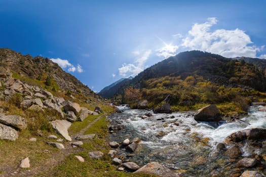 A wide shot captures a vibrant mountain river cascading over rocks, flanked by steep, rocky banks covered with patches of green and yellow vegetation.