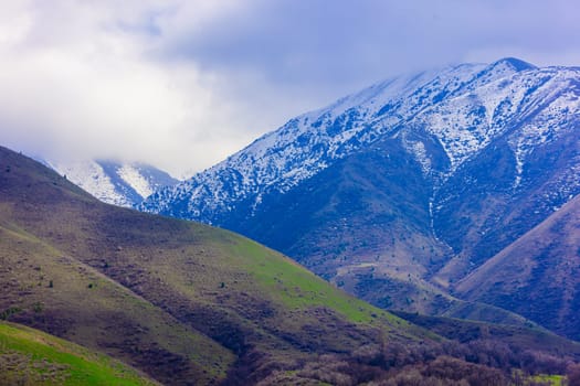 Snow-capped mountains in springtime Kyrgyzstan under a cloudy sky. Green and brown slopes.