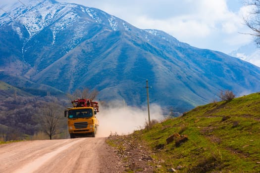 A bright yellow concrete boom pump truck travels along a dirt road, kicking up dust in a mountainous landscape.