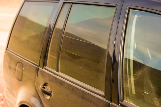 Rear view of a black vehicle covered in dust, with tinted windows reflecting the outdoor surroundings.