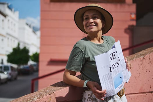 A delighted woman leans casually against a concrete railing, wearing a summer hat, holding a newspaper, and enjoying a sunny city day.