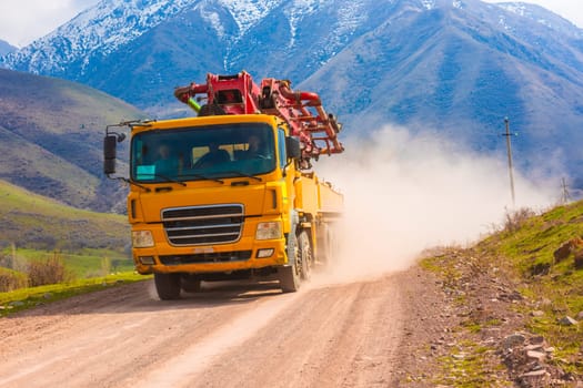 A bright yellow concrete pump truck drives towards the viewer on a dusty dirt road in a mountainous region.