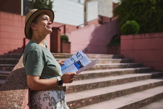 A cheerful woman in casual attire, enjoying a sunny day outdoors while reading a brochure. She exudes positivity as she leans against architectural stairs in a warm urban setting.