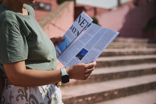 A person reads a newspaper outdoors in a sunny, tranquil location, enjoying leisure time on stairs with casual attire. The scene highlights urban outdoors and personal relaxation.