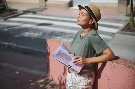 A stylish woman enjoys the sunny day on an urban sidewalk, wearing a hat, holding papers, and leaning against a reddish wall.