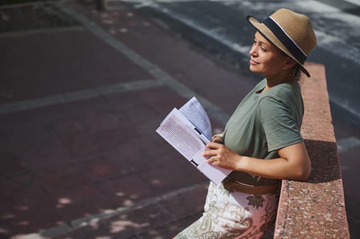 A content woman in a casual green shirt and straw hat enjoys reading a magazine while leaning against a wall on a sunny day outdoors.