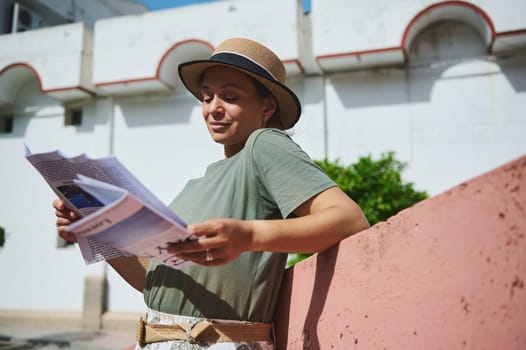 A young woman wearing a hat and casual attire reads a magazine outdoors on a sunny day, enjoying the warm weather and serene atmosphere.