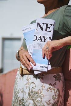 A stylishly dressed person holds a folded paper, featuring a printed text and design, while enjoying an outdoor setting in the sunlight.