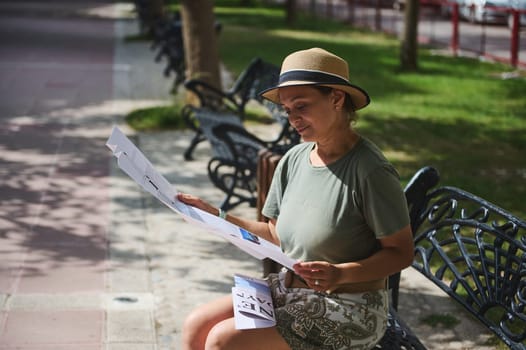 A woman sits on a park bench, enjoying the fresh air while reading a newspaper. She is wearing a summer hat, blending into the peaceful atmosphere of the open surroundings.
