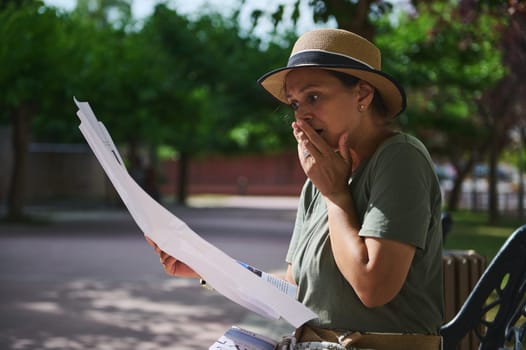 A woman seated in park surroundings displays a shocked reaction to a letter she reads, conveying emotional intensity.