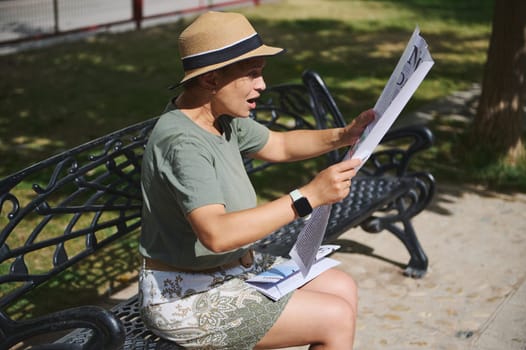 A woman enjoys reading a newspaper while sitting on a bench in a park. She appears engaged, wearing casual attire, under pleasant sunlight in a serene outdoor setting.