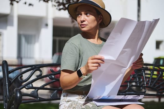 A thoughtful woman sits on a park bench while reading a large newspaper, enjoying the sunshine. She is wearing a straw hat and casual attire in a serene urban environment.
