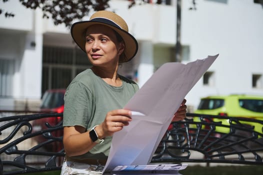 A woman wearing a green shirt and straw hat sits on a bench outdoors, examining a large paper under a bright sunny sky. The location provides a calm and engaging atmosphere.