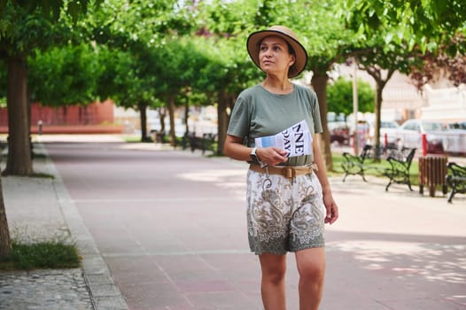 Casually dressed woman strolling along a park pathway lined with green trees and benches, holding a magazine. Bright, relaxed atmosphere ideal for leisure and outdoor concepts.