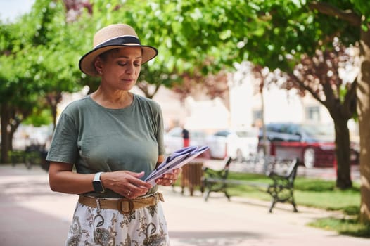 A woman enjoys a sunny day outdoors wearing a hat, reading a document while surrounded by trees and park benches, blending nature with urban tranquility.