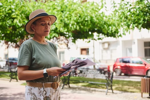 A woman wearing a hat and casual clothes reads a magazine in an urban park surrounded by trees and buildings.