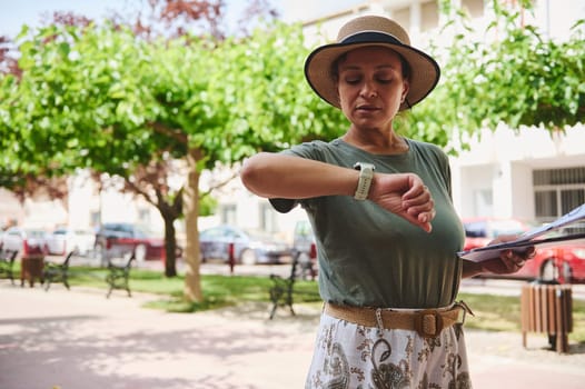 Focused woman in a summer hat outdoors checking her smartwatch while holding papers, conveying a feeling of modern technology and casual style.