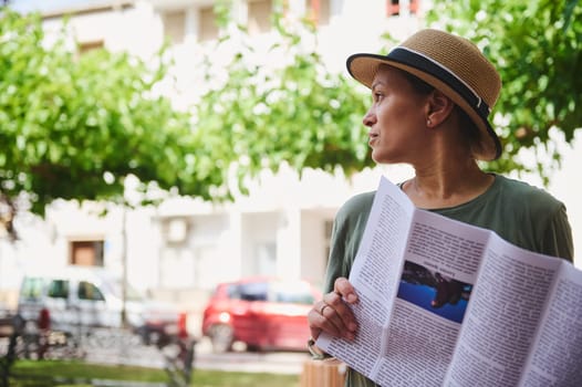 A woman wearing a hat with a magazine in hand, enjoying a quiet moment outdoors under the trees, surrounded by natural light and urban elements.