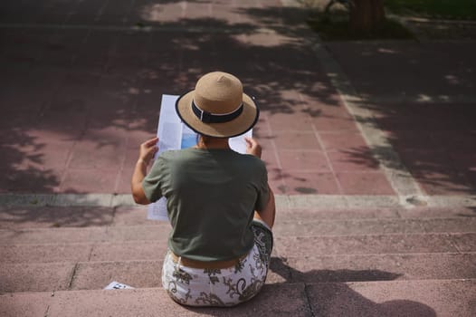 A woman sits on outdoor steps wearing a straw hat and holding papers. The sunny setting suggests relaxation and focus in an urban park environment.