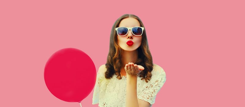 Portrait of beautiful young woman blowing her lips sending sweet air kiss with balloon on pink background