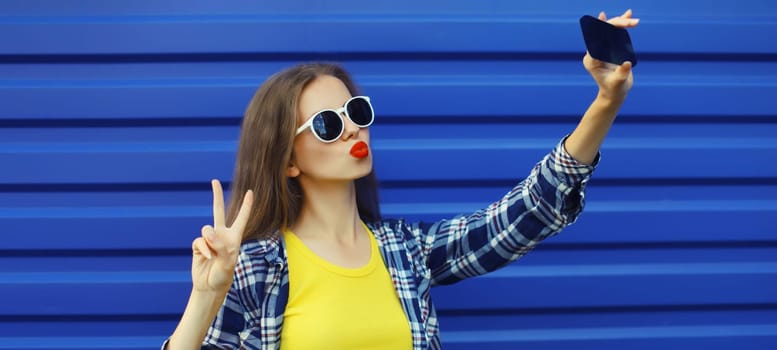 Summer cheerful stylish young woman taking selfie with smartphone, cool modern girl blowing a kiss in glasses on blue background