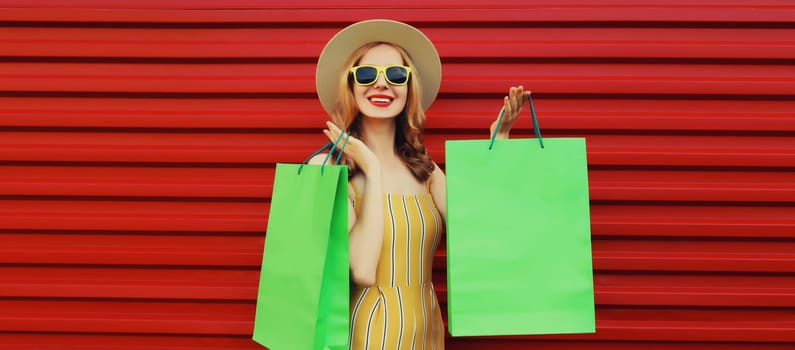 Fashion, purchase. Portrait of stylish beautiful happy young woman posing with white shopping bags, girl wear summer hat, dress standing on bright red background