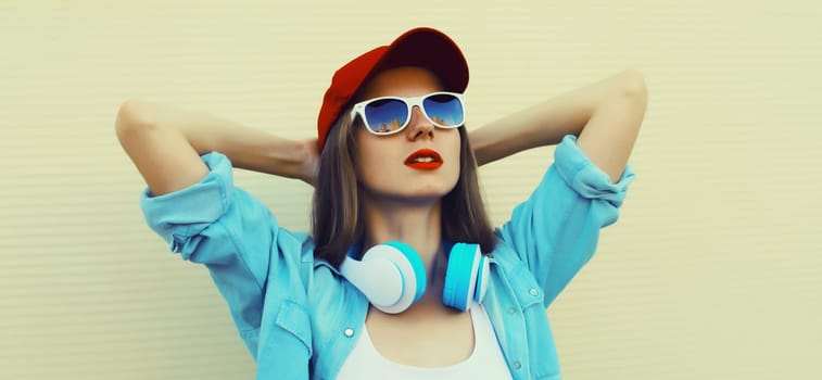 Stylish modern young woman listening to music with headphones in baseball cap, casual clothing on white background