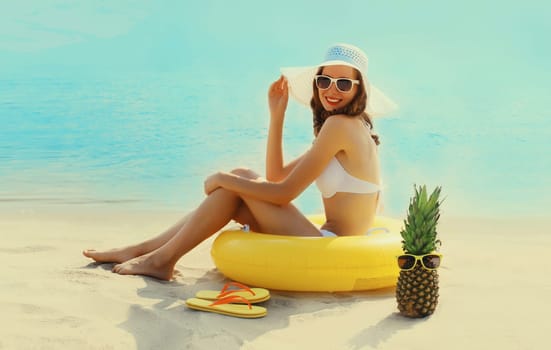 Summer vacation, tourism, happy smiling woman with swimming inflatable ring, tourist hat, pineapple, fruits, sunglasses, sitting on the sand on the beach on sea coast background