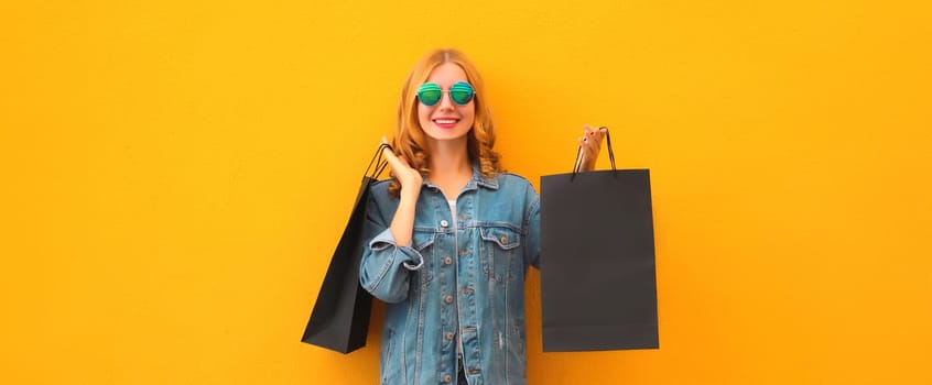 Shopping day, stylish beautiful happy smiling young woman posing with black shopping bags wearing denim clothing on yellow studio background