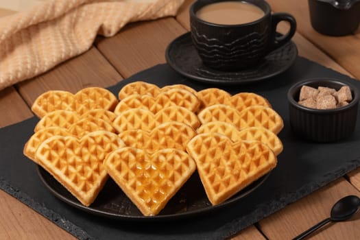 Heart-shaped waffles are elegantly arranged on a black plate, accompanied by a cup of steaming coffee and a small bowl of sugar cubes, creating a warm and inviting atmosphere.