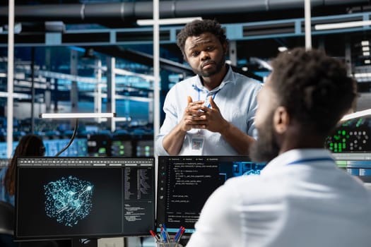 Server farm manager supervising engineer looking at data analysis graph to reduce failures. Data center team leader overseeing worker looking at AI visualization dashboard to pinpoint gear glitches