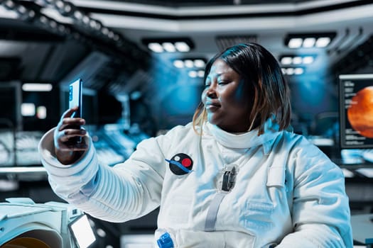 Female astronaut doing videocall, talking with family, presenting space station. African american woman in spaceship using smartphone to recording herself sharing mission progress with loved ones