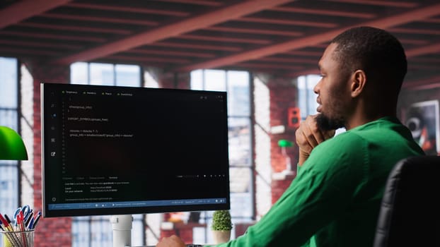 Black technician in home office looks at UI on computer monitor, typing and executing programming code. IT worker using coding application to write and test code in modern apartment. Camera A.