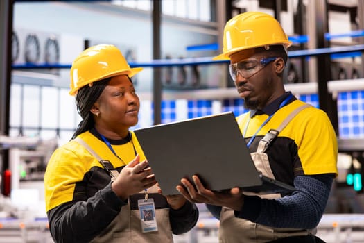 In smart manufacturing plant, african american engineers supervise industrial automation system using laptop. The automated factory features conveyor belts, robotic arms and optimized assembly lines.