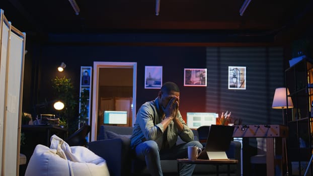 Tired black man failing some tasks and struggling to focus at home, dealing with burnout after doing overtime. Overworked fatigued person closes the laptop and taking a break. Camera A.