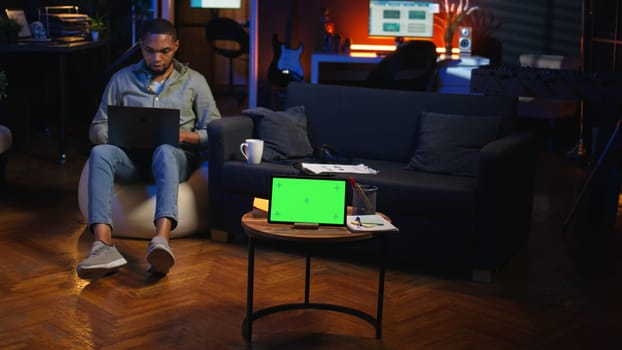 African american male freelancer enjoys free time next to chroma key on the bean bag, uses device to watch a podcast with headphones. Young guy relaxing remotely in the living room. Camera A.