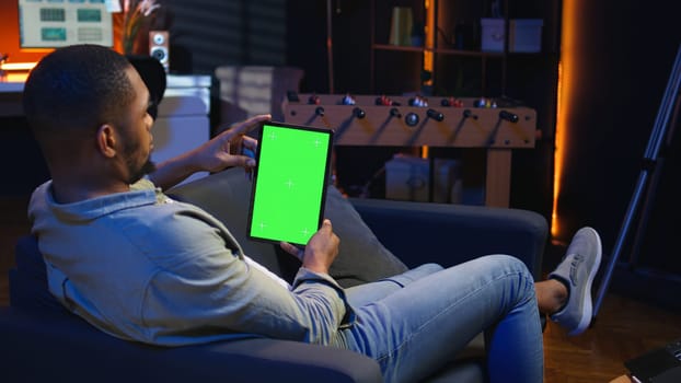Black remote worker looking at green screen on his device at home, relaxing in the living room and solving freelancing tasks. Guy being casual in trendy apartment, teleworking. Camera A.